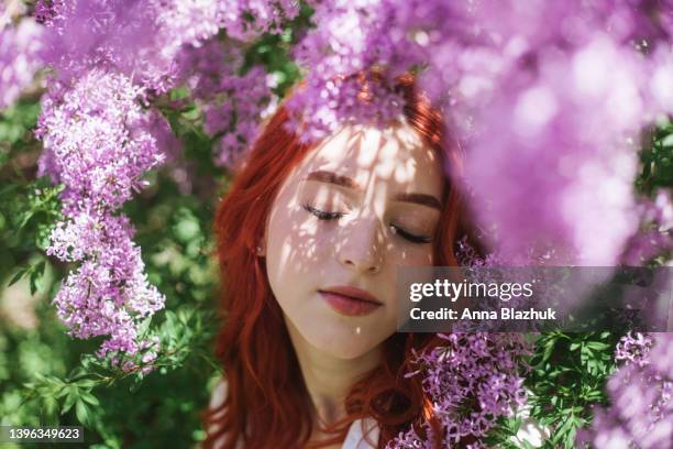 young woman with red long curly hair in white shirt sitting near blossoming purple lilac bush in spring park in sunny day. female romantic portrait. - purple lilac stock pictures, royalty-free photos & images