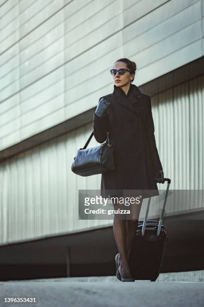 young elegant woman walks down the sidewalk next to an office building and pulls small luggage on wheels - woman standing looking down stock pictures, royalty-free photos & images