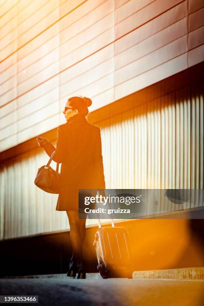 young elegant woman walks away down the sidewalk next to an office building and pulls small luggage on wheels - small office building stock pictures, royalty-free photos & images