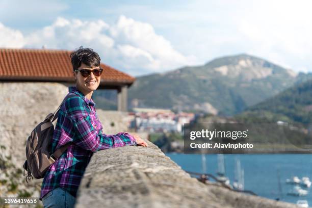 portrait of smiling young woman with short hair and sunglasses with beautiful landscape lighthouse and sea port - getönte sonnenbrille stock-fotos und bilder
