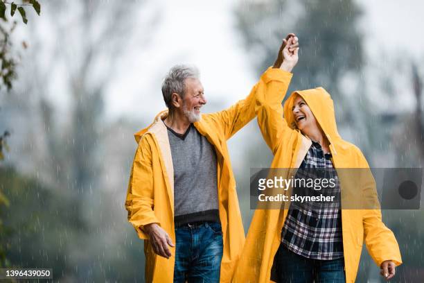 happy mature couple in raincoats dancing on rain at the park. - people rain happy stockfoto's en -beelden