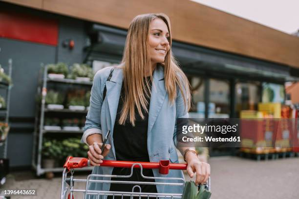 smiling woman with smart phone and shopping cart standing in front of store - shopping trolleys stock pictures, royalty-free photos & images
