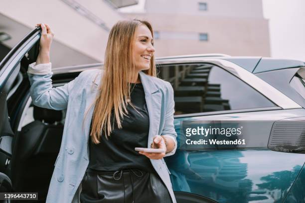 happy woman with mobile phone standing by car door - personenauto stock-fotos und bilder