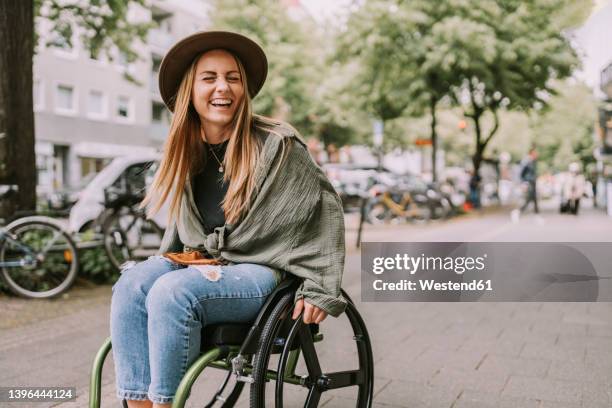 cheerful young woman sitting in wheelchair at sidewalk - junge frau im rollstuhl stock-fotos und bilder