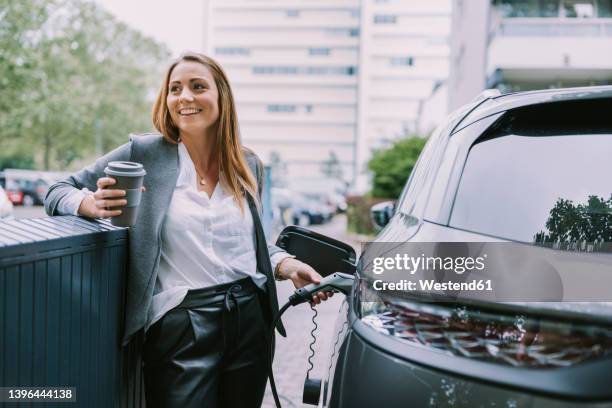 happy woman with disposable coffee cup charging car at station - electric vehicle ストックフォトと画像