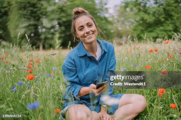 happy woman with smart phone sitting amidst plants in meadow - chignon fotografías e imágenes de stock