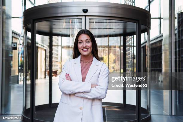 smiling businesswoman with arms crossed standing in front of revolving door - revolving door stock pictures, royalty-free photos & images