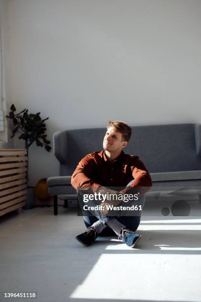 smiling man sitting on floor in front of sofa in living room at home - sitting on floor fotografías e imágenes de stock