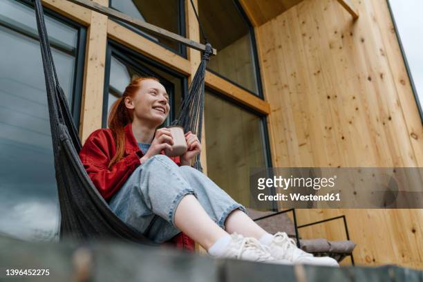 smiling woman with coffee cup in hammock chair at deck - rote haare stock-fotos und bilder