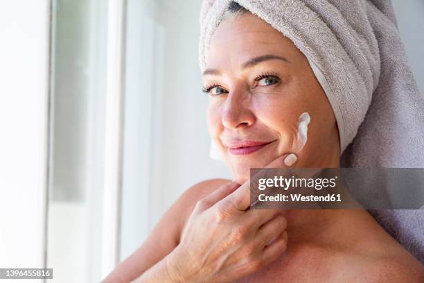 smiling mature woman applying moisturizer on face at home - cream for face stock pictures, royalty-free photos & images