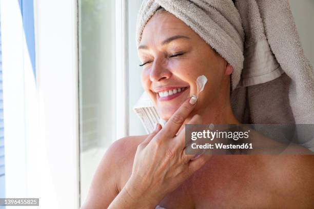 happy woman with eyes closed applying moisturizer on face at home - gezichtscrème stockfoto's en -beelden