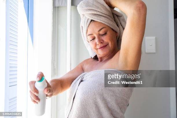 smiling woman wearing towel spraying deodorant on body at home - female armpits stock pictures, royalty-free photos & images