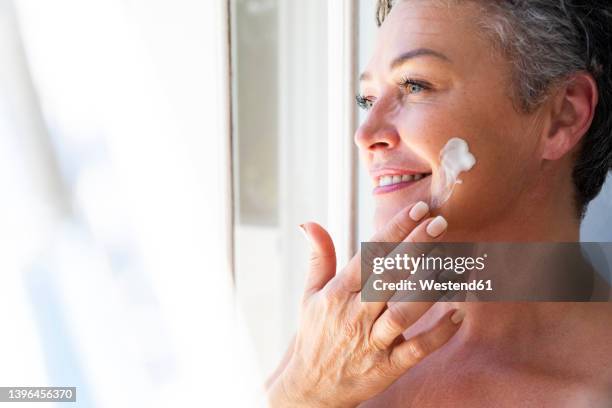 smiling mature woman applying moisturizer cream on face - beauty treatment fotografías e imágenes de stock