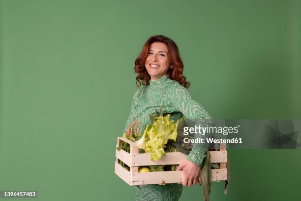 smiling woman holding wooden crate with vegetables against green background - woman hold box imagens e fotografias de stock