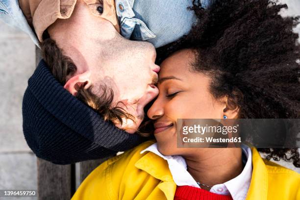 man with eyes closed kissing girlfriend on forehead - forehead stock-fotos und bilder