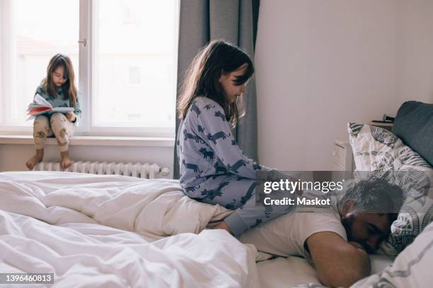 girl sitting on father's back sleeping on bed while sister reading book at window - child in bed clothed stock-fotos und bilder