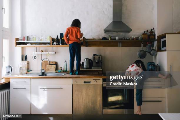 rear view of girl searching on shelf while sister climbing kitchen counter - clambering stock pictures, royalty-free photos & images