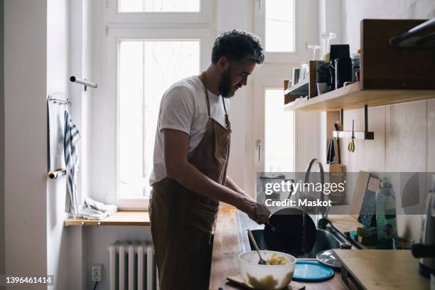 side view of man wearing apron washing dishes in kitchen at home - afwas doen stockfoto's en -beelden