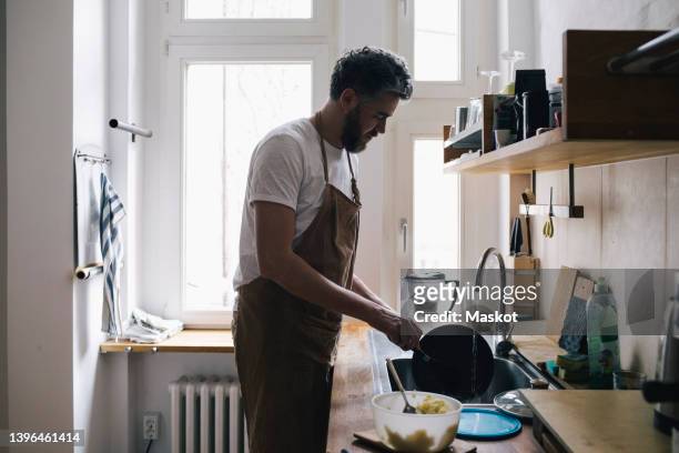 side view of man wearing apron washing dishes in kitchen at home - men housework stock pictures, royalty-free photos & images