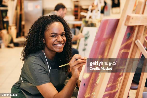 portrait of smiling mature woman with paintbrush sitting by easel in art class - blank canvas stockfoto's en -beelden