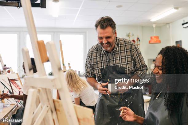 smiling mature male tutor looking at artist's canvas by student sitting in art class - art class stockfoto's en -beelden