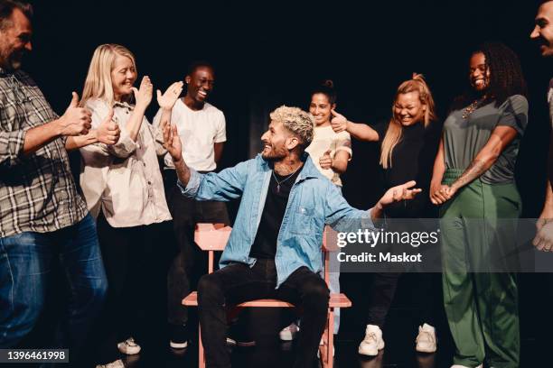 happy young man sitting on chair amidst multiracial stage performers in class - répétition de spectacle photos et images de collection