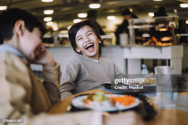 boy laughing while sitting with friend during lunch break at school - cafeteria stockfoto's en -beelden