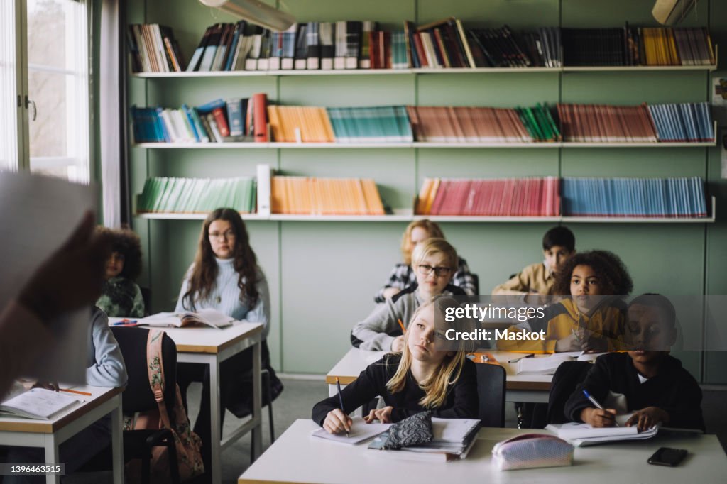Diverse students writing while listening to teacher during lecture in classroom