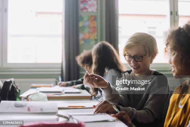 smiling friends playing while sitting at desk in classroom - brugklas stockfoto's en -beelden