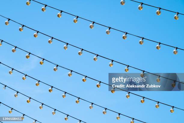 a garland of light bulbs in a row hangs outdoors. decorative lanterns, against a blue sky background. copy space. - lightbulbs in a row stock pictures, royalty-free photos & images
