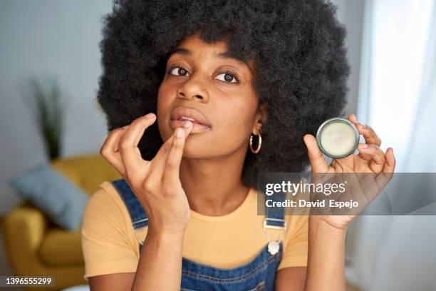 young afro woman applying lip balm on her lips while doing her make-up at home. - shiny lips stock pictures, royalty-free photos & images