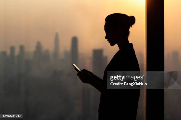 silhouette of young woman using smartphone next to window with cityscape - surveillance stockfoto's en -beelden