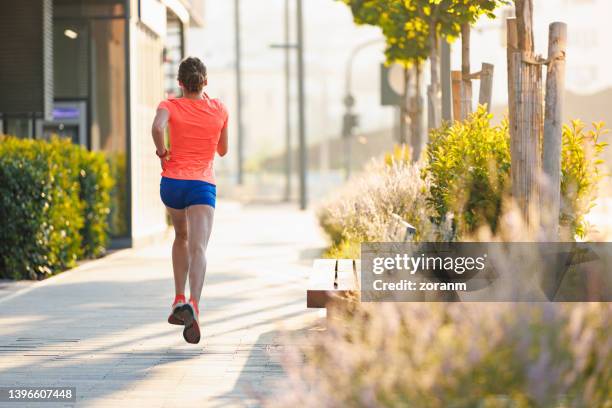 vista posteriore sulla giovane donna in pantaloncini da corsa a mezz'aria, jogging sul marciapiede - running shorts foto e immagini stock