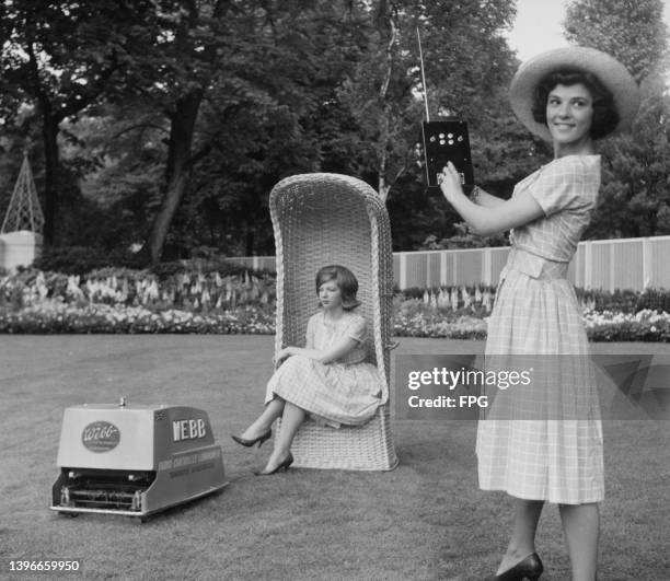 Two women, one sitting in a wicker garden chair while the other stands with the controller of the Webb radio-controlled electric lawnmower in the...