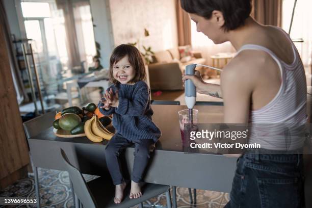view of a single mom making a fruit cocktail for her daughter. small girl sitting on a table and smiling. - family small kitchen stock pictures, royalty-free photos & images