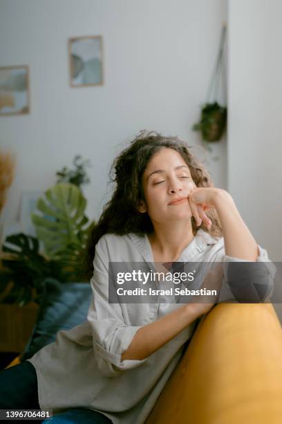 young white curly-haired woman with her eyes closed as she relaxes sitting on the sofa at home. - gente tranquila fotografías e imágenes de stock