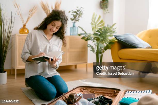 horizontal view of a young woman making a checklist with her notebook while preparing her travel suitcase to go on vacation. - packing travel stock-fotos und bilder