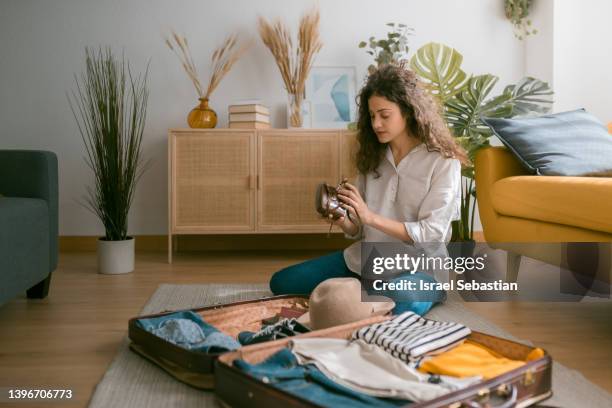 horizontal view of a young curly-haired caucasian woman sitting on the floor stowing her analog camera in her suitcase for a vacation trip to the salon. - suitcase packing stockfoto's en -beelden