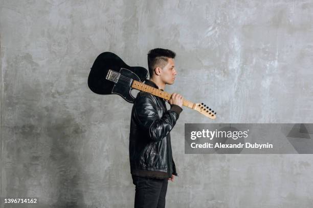 a handsome brunette man with a guitar in his hand and on his shoulder walks against the background of a concrete wall - pop musician stock pictures, royalty-free photos & images