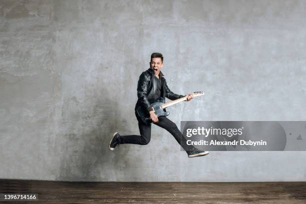 emotional portrait of a rock guitarist jumping with a guitar in his hands against the background of a concrete wall with copy space. happy guy artist have fun in studio with musical instrument. - asian singer stock pictures, royalty-free photos & images