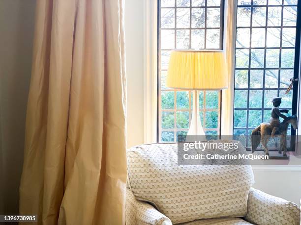 upholstered antique low back wing chair in corner of living room in front of illuminated lamp & beside curtain panel - american family fine art foto e immagini stock