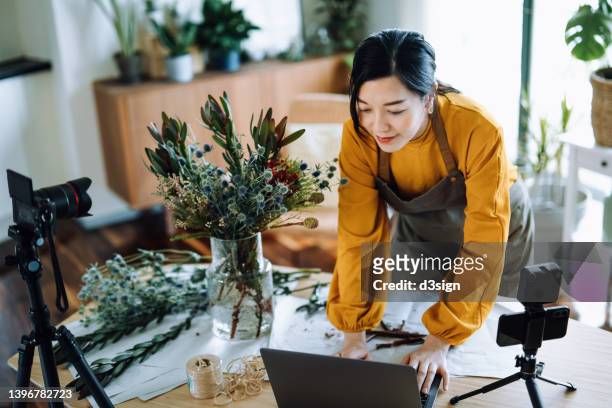professional young asian female florist working on laptop while making vlog session on flower bouquet arrangement class with laptop and camera at home. self-employment. online classes. small business concept - florista stock-fotos und bilder