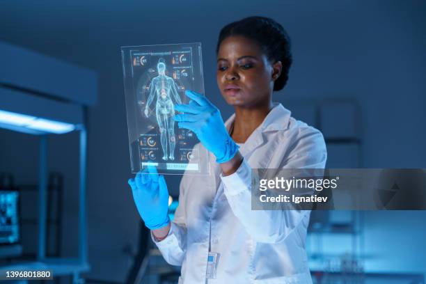 a young concentrated african - american scientist in a lab coat and protective gloves works on a small hud or graphic display with the image of a human body - head mounted display stockfoto's en -beelden