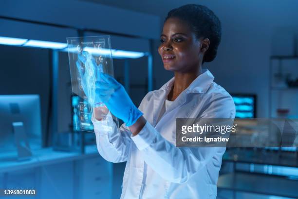 a young smiling african - american scientist in a lab coat and protective gloves works on a small hud or graphic display in front of her, standing in a modern lab - head mounted display stockfoto's en -beelden