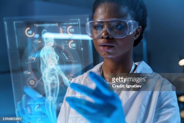 close-up of a young concentrated african - american scientist in a lab coat and protective gloves working on a small hud or graphic display in front of her with the image of a human body - head mounted display stockfoto's en -beelden