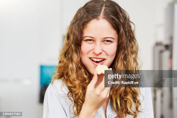 beautiful young woman with long curly hair takes a bite out of a slice of apple in her kitchen - mastigar imagens e fotografias de stock