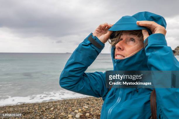 young woman looking up at stormy sky, pulls on protective hood over head - waterproof clothing stock pictures, royalty-free photos & images