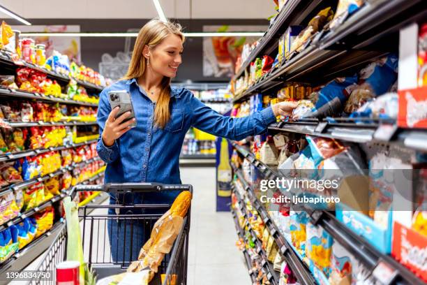 bella giovane donna cliente compra generi alimentari utilizzando uno smartphone che cammina con un carrello al supermercato. - wagon foto e immagini stock