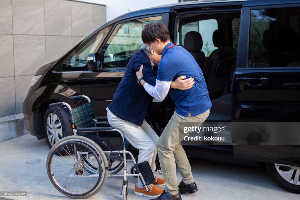 A senior man helped by a young male carer out of wheelchair and into car