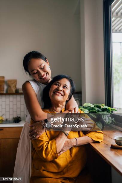porträt einer asiatischen zweierfamilie, mutter und tochter, die in einer küche sitzt, tochter, die ihre mutter mit viel liebe und hingabe umarmt. - happy family asian stock-fotos und bilder